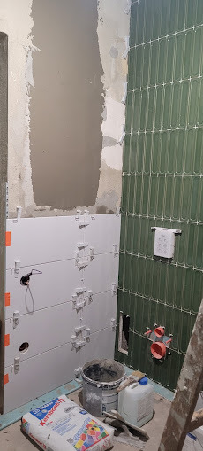 Wnętrze łazienki w trakcie prac. Na ścianach białe i zielone płytki, widoczne krzyżyki dystansowe. Na podłodze wiadro z zaprawą i worek kleju.