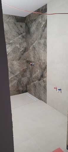 Wnętrze łazienki w trakcie wykańczania: marmurowe płytki na ścianie, biała podłoga z odpływem liniowym, widoczne instalacje wodne. Surowy stan, nowoczesny design.