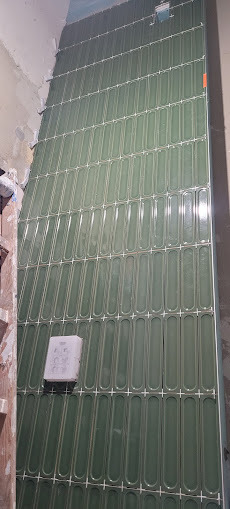 Ściana w trakcie wykańczania z podłużnymi, zielonymi płytkami, widoczne krzyżyki dystansowe i niedokończone fragmenty ściany. Ujęcie z dołu.