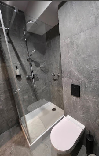 Nowoczesna łazienka z kabiną prysznicową typu walk-in, wykończona szarymi płytkami imitującymi marmur. Widoczna toaleta i akcesoria łazienkowe w minimalistycznym stylu.