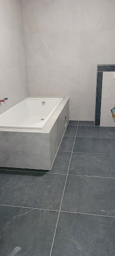 Minimalistyczna łazienka z wanną obudowaną szarą płytką i ciemną podłogą. Ściany wykończone jasnymi, dużymi płytami. Widok na narożnik pomieszczenia.