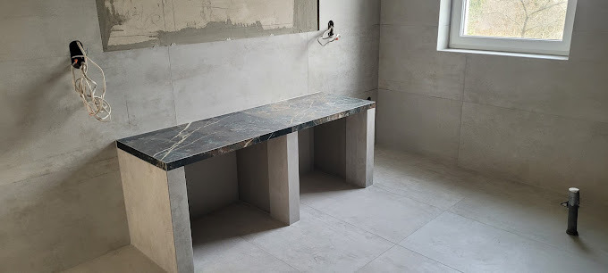 Wykończona łazienka z szarą glazurą i blatem imitującym marmur. Widoczne niedokończone instalacje elektryczne i okno. Minimalistyczny design.