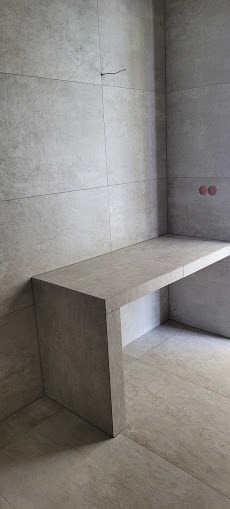 Minimalistyczna łazienka z szarymi płytkami imitującymi beton, zabudowa umywalkowa z tego samego materiału. Widoczne gniazdka elektryczne i wystający kabel.