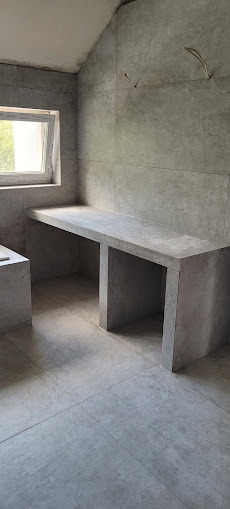 Szara łazienka z blatem z płytek, widok na okno i fragment wanny. Minimalistyczny design, płytki imitujące beton. Widoczne przewody elektryczne.