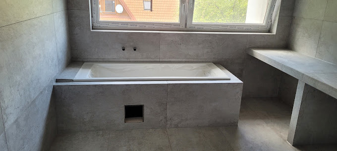 Nowoczesna łazienka z wanną obudowaną szarą płytką, półką i oknem dachowym. Minimalistyczny design, szare płytki na ścianach i podłodze. Widok na fragment dachu za oknem.