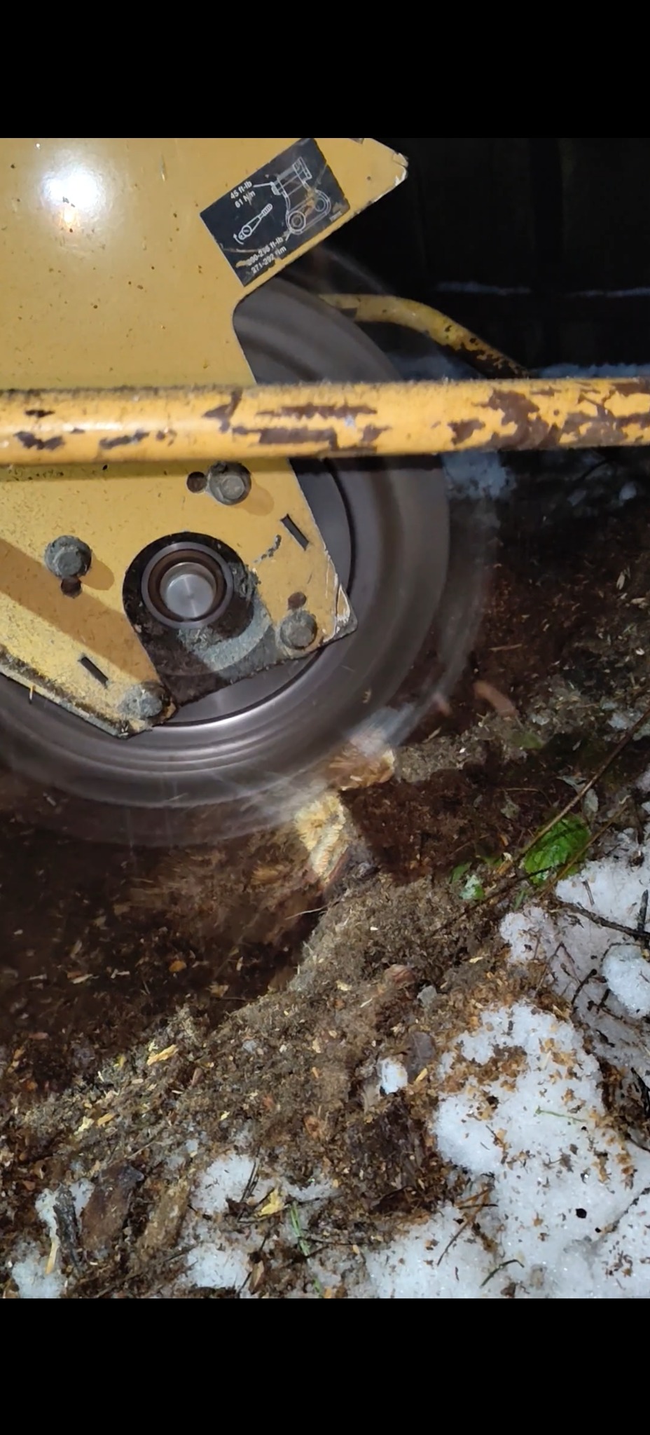 Żółta frezarka do pni w akcji, usuwająca pień drzewa. Wokół widać wióry i resztki drewna, a także śnieg. Maszyna w trakcie pracy.