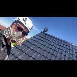 Specjaliści od Renowacji,Wysokości i Wycinek 66-400 - Pracownik w białym kasku i masce ochronnej, z okularami, aplikuje powłokę na ciemne, błyszczące dachówki, widoczne świeże wykończenie i błękitne niebo.
