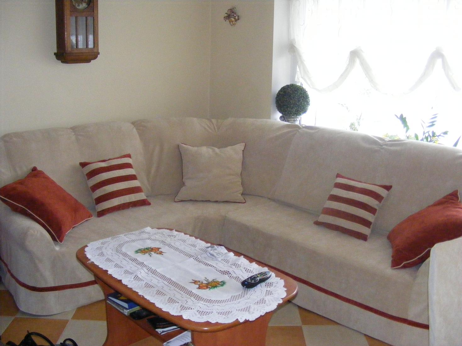 Beżowy narożnik z bordową lamówką i dekoracyjnymi poduszkami w salonie, stolik kawowy z haftowanym obrusem, w tle okno z firanką i dekoracyjna kula z roślin.