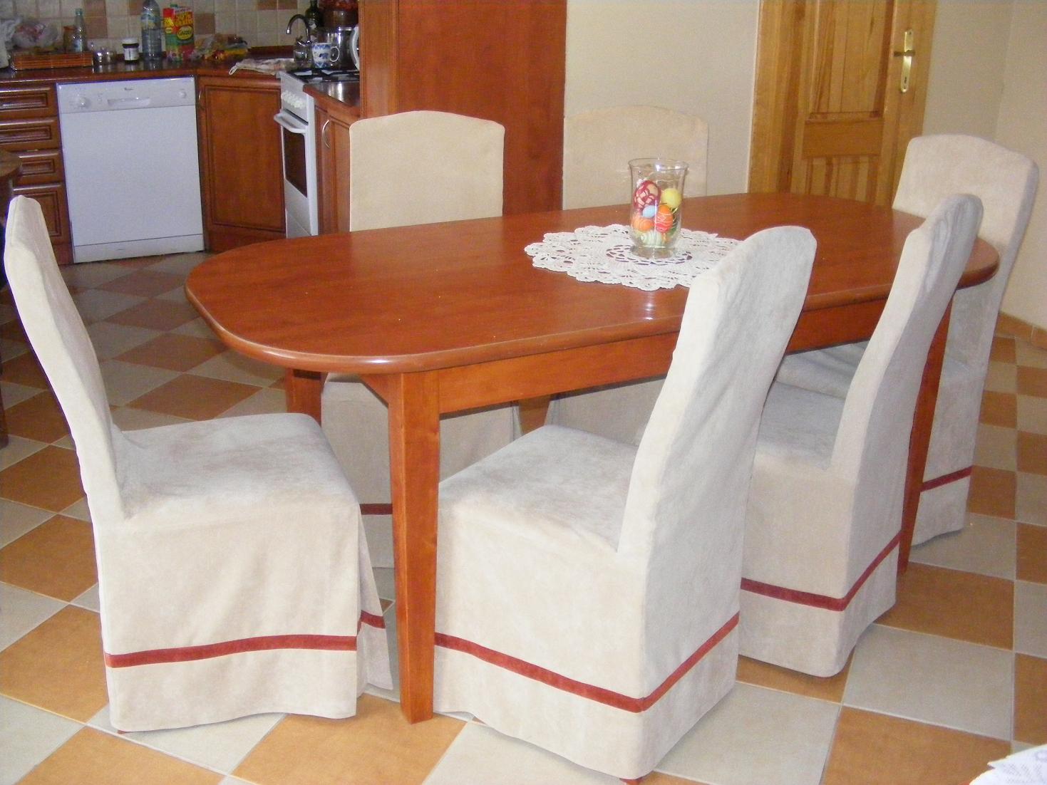 Jadalnia z owalnym, drewnianym stołem nakrytym białą serwetą i szklanym wazonem z dekoracjami, otoczonym krzesłami z beżowymi pokrowcami z bordową lamówką.