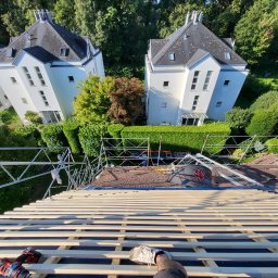 Przemysław Gryciak Modern House - Montaż łat dachowych na tle zieleni i białych domów. Widok z góry, narzędzia pracy i fragment buta w kadrze. Prace dekarskie w toku, z użyciem wkrętarki.