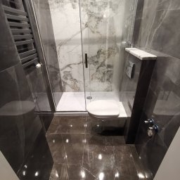 Nowoczesna łazienka z kabiną prysznicową typu walk-in, marmurowymi płytkami w odcieniach szarości i białym, podwieszanym WC. Widoczny grzejnik drabinkowy.