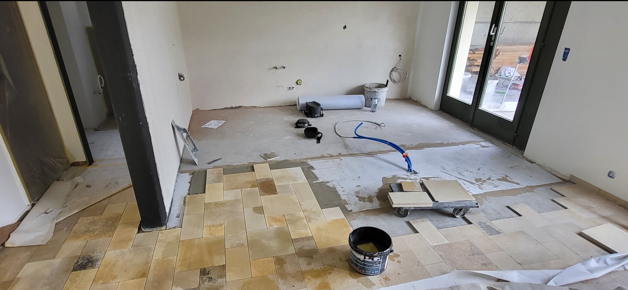 Układanie beżowej terakoty na podłodze w trakcie remontu. Widoczne narzędzia glazurnicze, wiadro z klejem i fragmenty niezakończonej podłogi. Jasne wnętrze z dużym oknem.