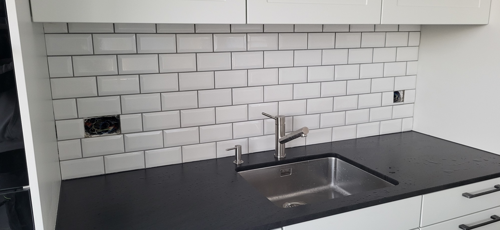 Białe, błyszczące płytki typu metro z ciemną fugą nad czarnym blatem kuchennym ze stalowym zlewem i baterią. Widoczne gniazdka elektryczne bez osprzętu.