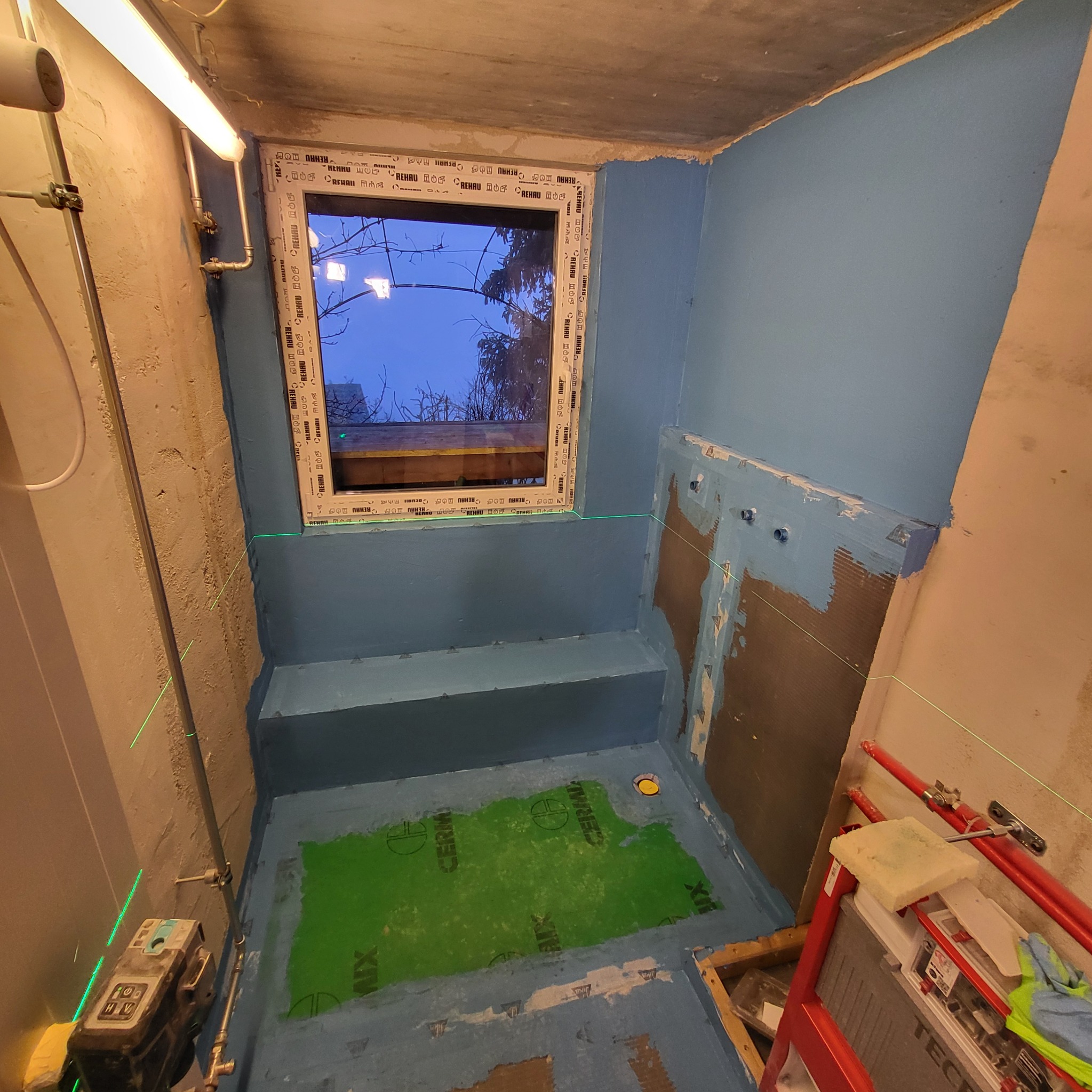 Hydroizolacja w łazience w trakcie remontu. Widoczne narzędzia, okno z widokiem na zewnątrz i zielona mata hydroizolacyjna na podłodze. Laser krzyżowy wyznacza poziom.