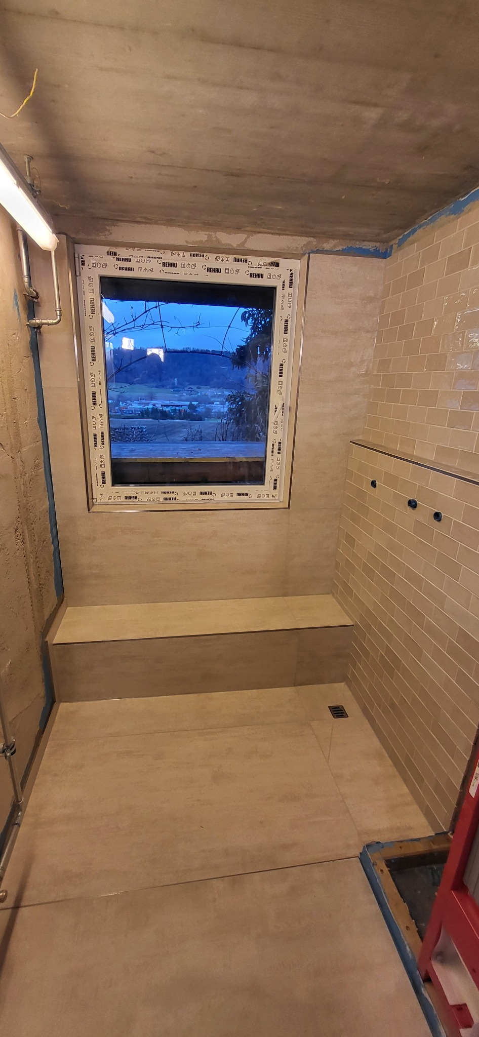 Wykończona łazienka z oknem i beżowymi płytkami, widok na krajobraz za oknem. Ukończona zabudowa z płytek tworząca siedzisko. Kratka ściekowa w podłodze.