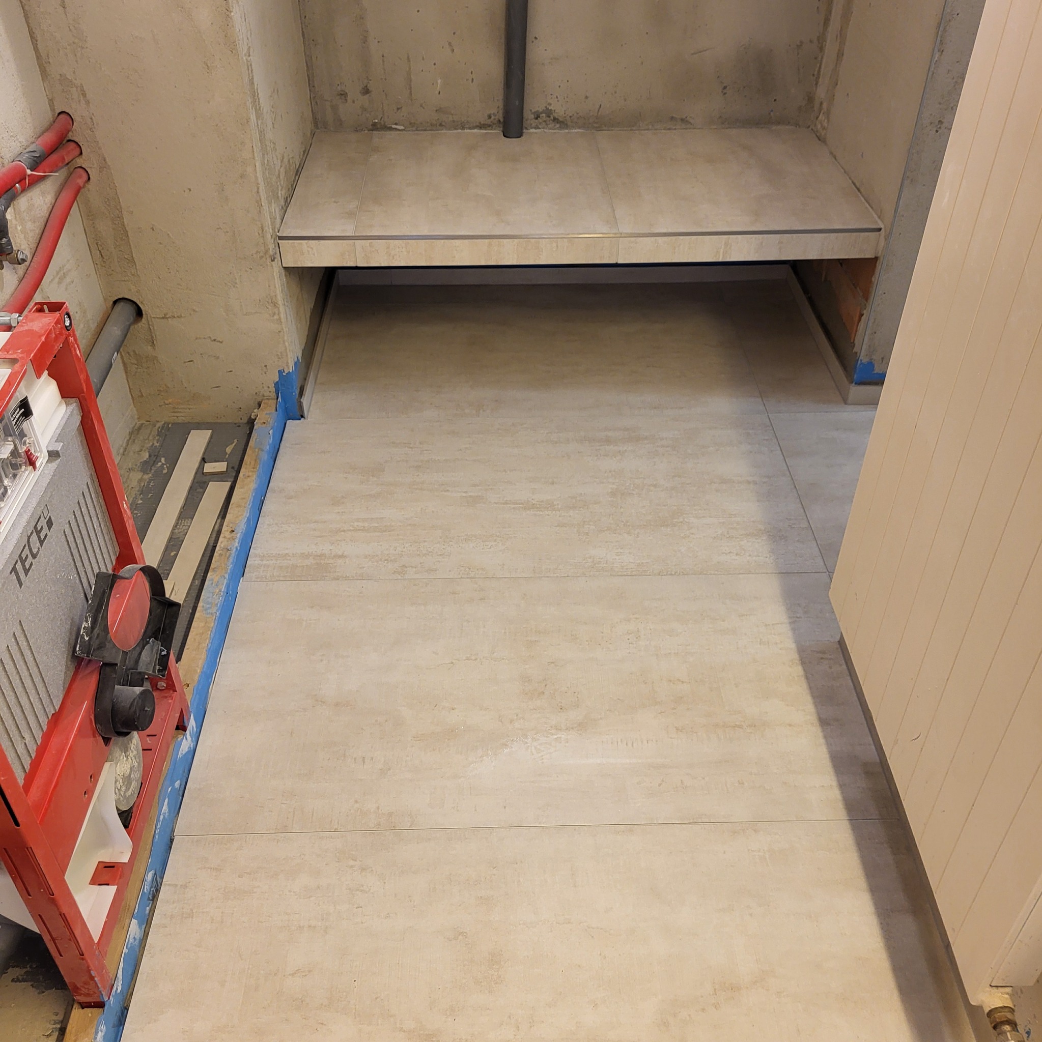 Ułożone beżowe płytki podłogowe w łazience, widoczny stelaż WC Tece i fragment ściany z instalacją hydrauliczną. Minimalistyczny design, surowe wykończenie.