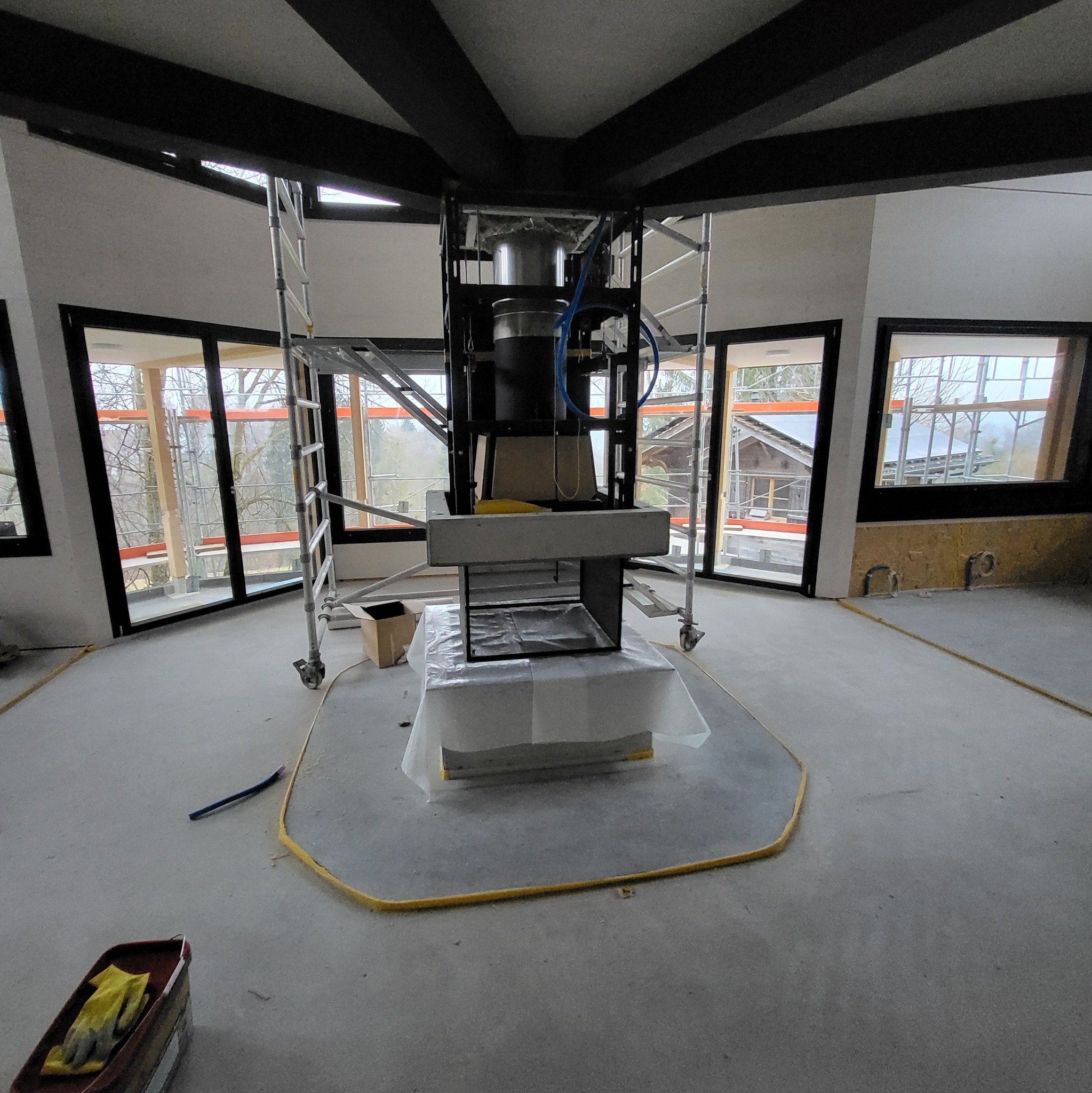 Wnętrze w trakcie prac wykończeniowych: surowy beton, rusztowania, okna z widokiem na zewnątrz. Przygotowanie do montażu kominka w centralnej części pomieszczenia.