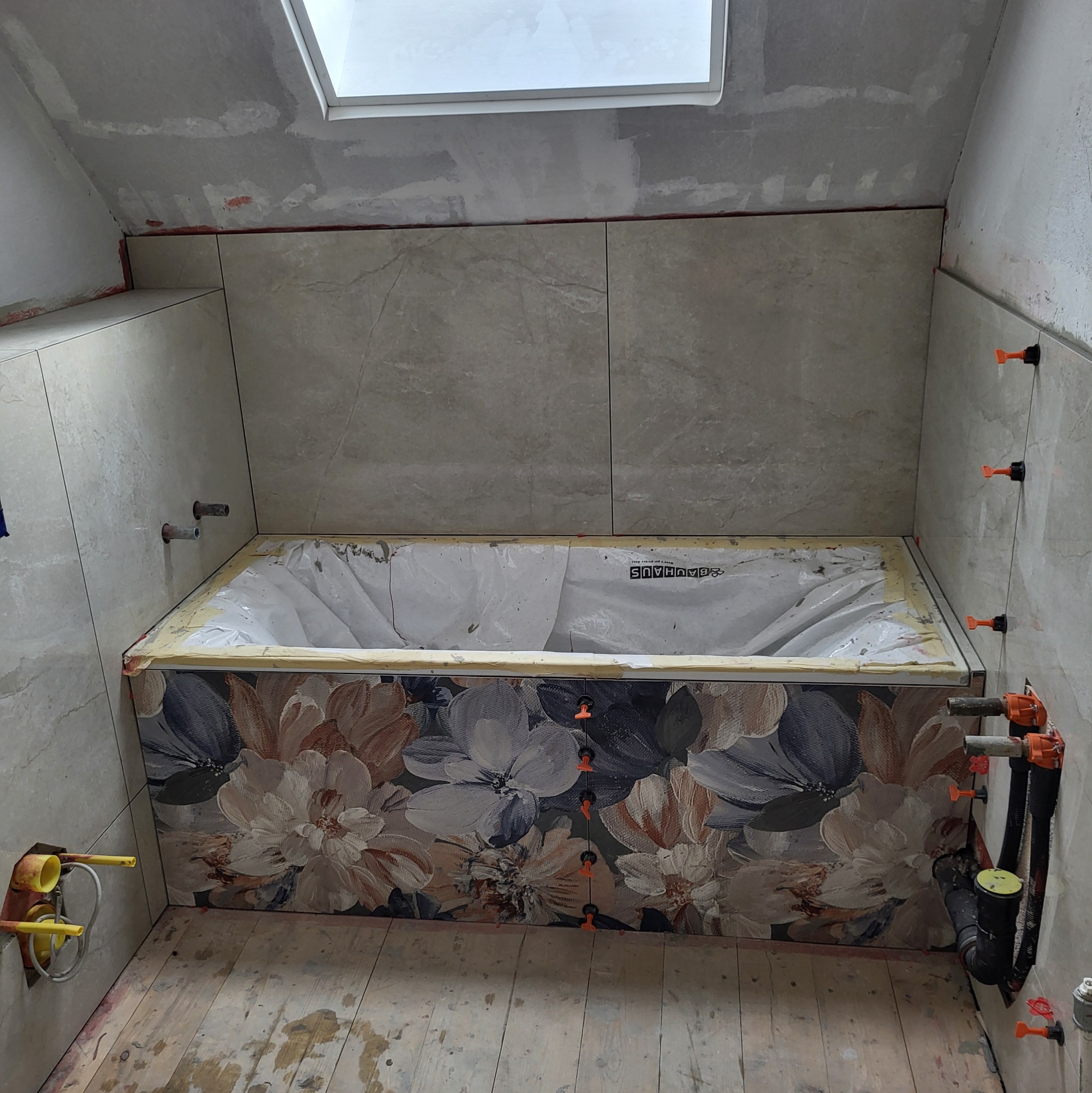 Montaż płytek z motywem kwiatowym wokół wanny w łazience. Widoczne narzędzia glazurnicze i fragment okna dachowego. Prace w toku.