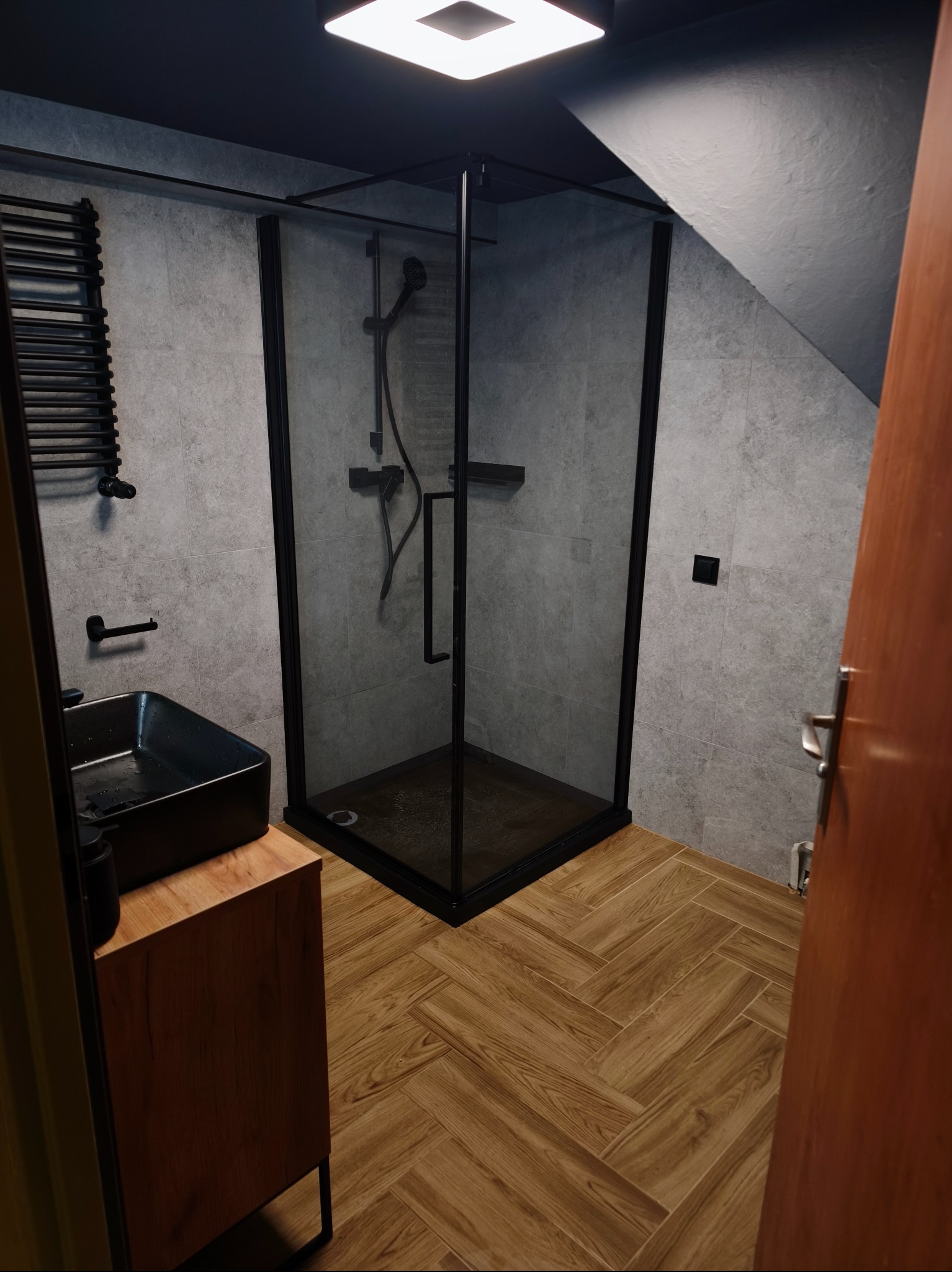 Nowoczesna łazienka z kabiną prysznicową typu walk-in z czarnymi ramami, umywalką nablatową i grzejnikiem drabinkowym na tle szarych płytek i drewnopodobnej podłogi.