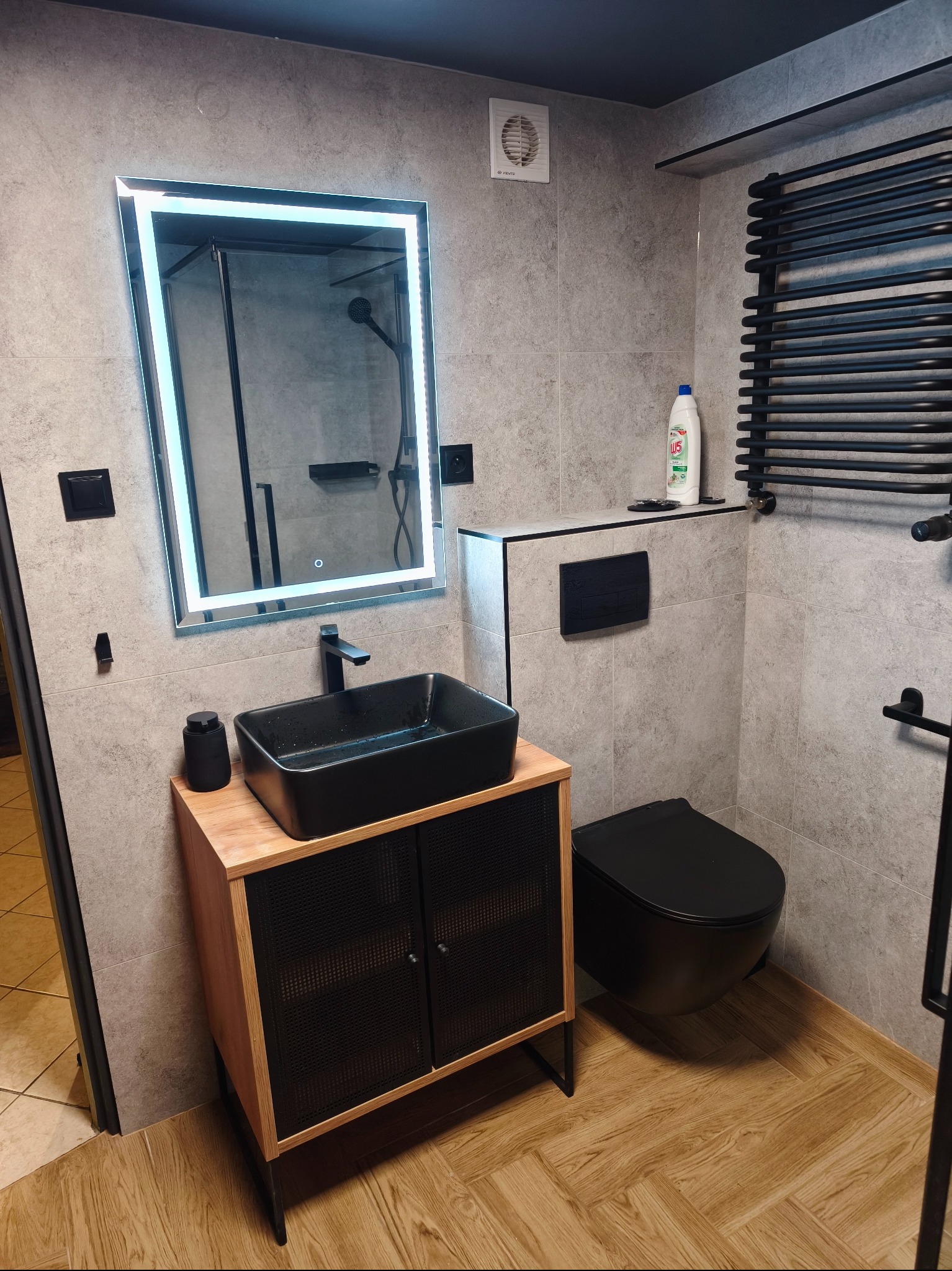 Nowoczesna łazienka w szarościach z czarnymi akcentami: umywalka na szafce z drewnianym blatem, podświetlane lustro, wisząca toaleta i grzejnik drabinkowy.