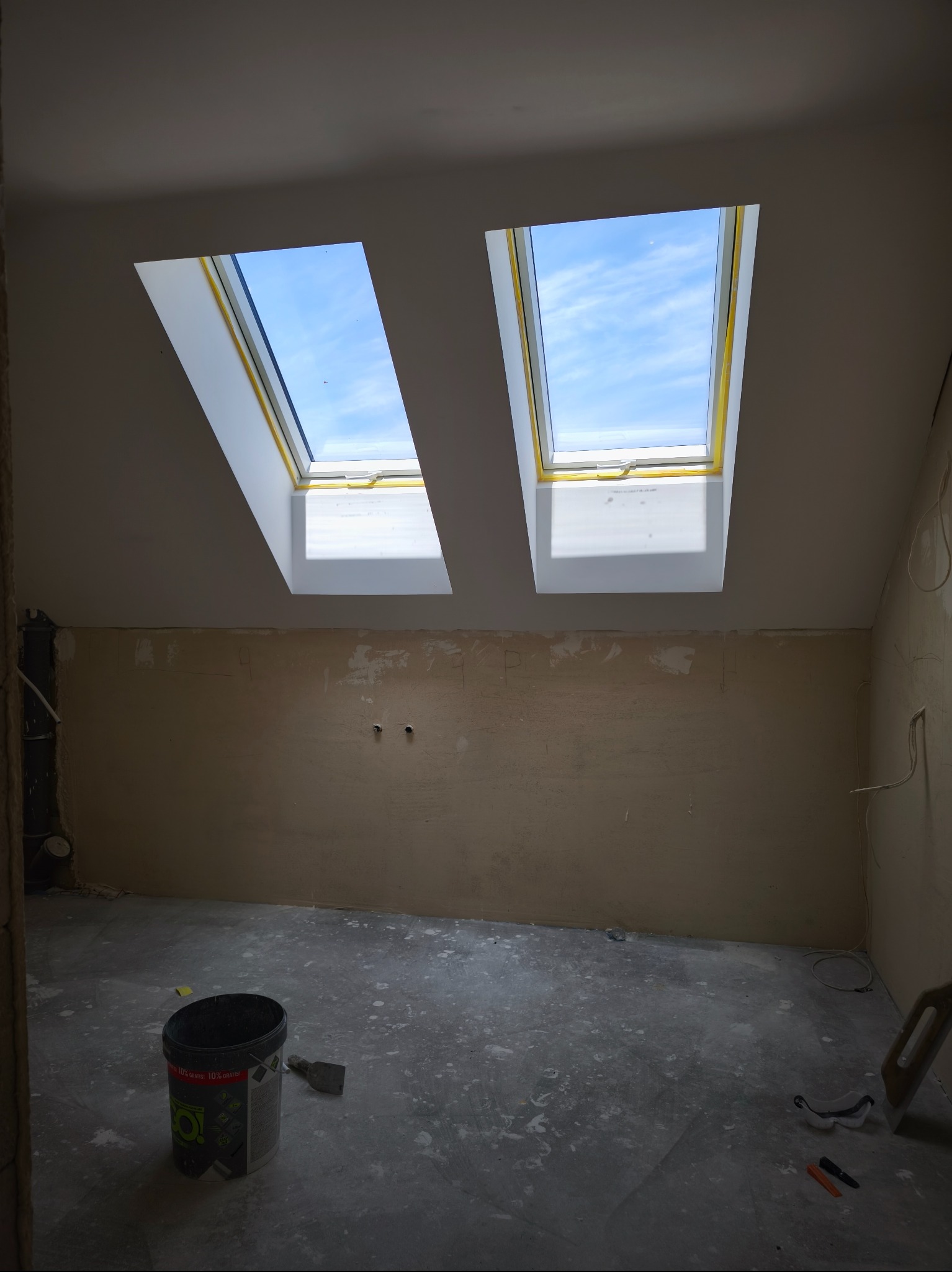 Pokój na poddaszu w trakcie remontu: ściany przygotowane do szpachlowania, widoczne dwa okna dachowe z widokiem na błękitne niebo, wiadro z farbą i narzędzia.