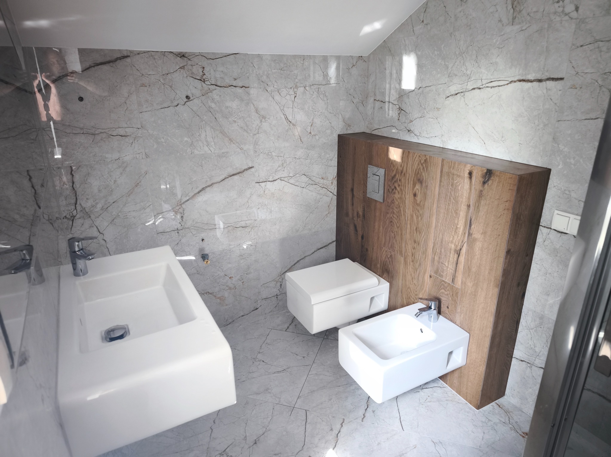 Nowoczesna łazienka z marmurowymi płytkami, wiszącą toaletą i bidetem oraz umywalką. Drewniana zabudowa stelaża WC. Minimalistyczny design, jasne wnętrze.