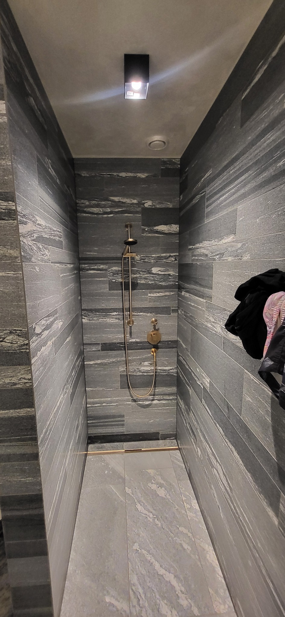 Nowoczesna kabina prysznicowa z szarymi płytkami imitującymi kamień, złotą armaturą i minimalistycznym oświetleniem. Widok na wnętrze kabiny.