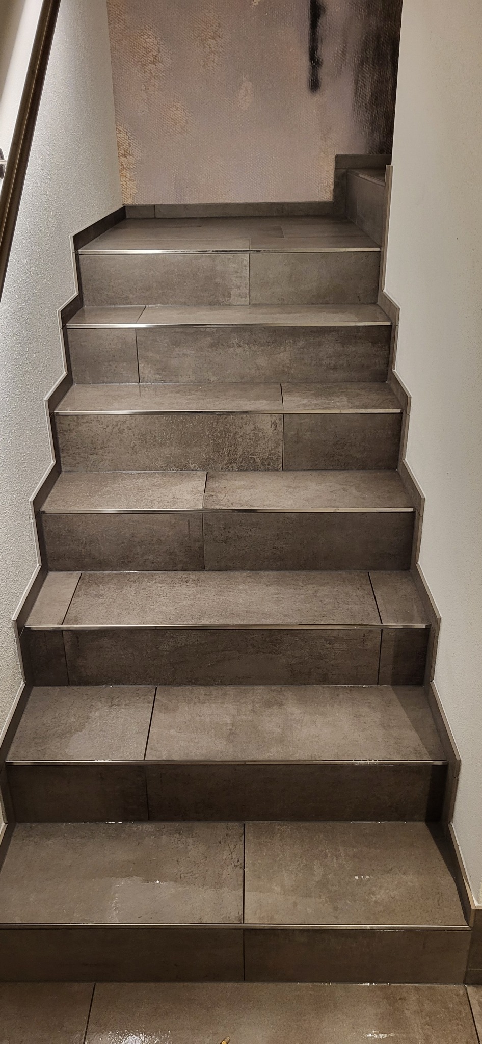 Wykończone schody z szarych płytek z metalowymi listwami, widok z góry. Płytki ułożone z przesunięciem, ściany białe i beżowe. Widoczne detale wykończenia.