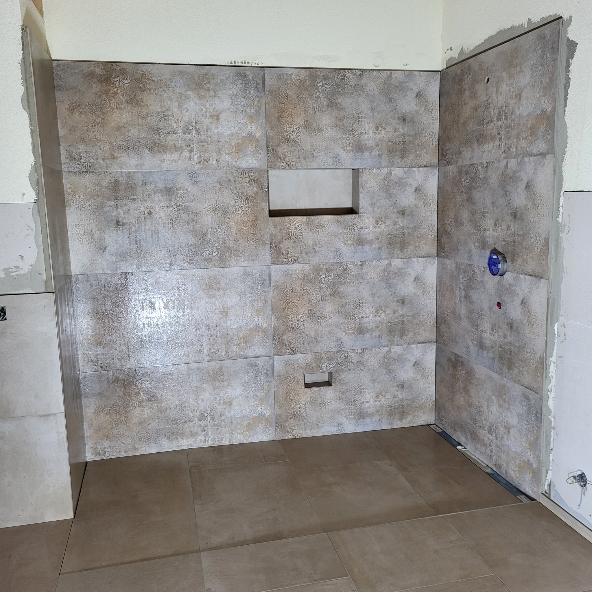 Wykończona ściana i podłoga w łazience z beżowych płytek z wzorem, widoczne otwory na armaturę i wnęki. Prace glazurnicze w trakcie realizacji.