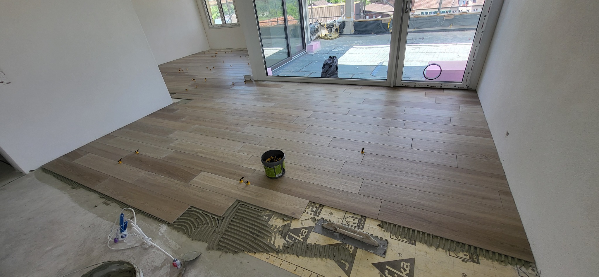 Układanie podłogi z płytek imitujących drewno w jasnym wnętrzu. Widoczne narzędzia glazurnicze i poziomice. Częściowo ułożona podłoga z fugami i elementami dystansowymi.