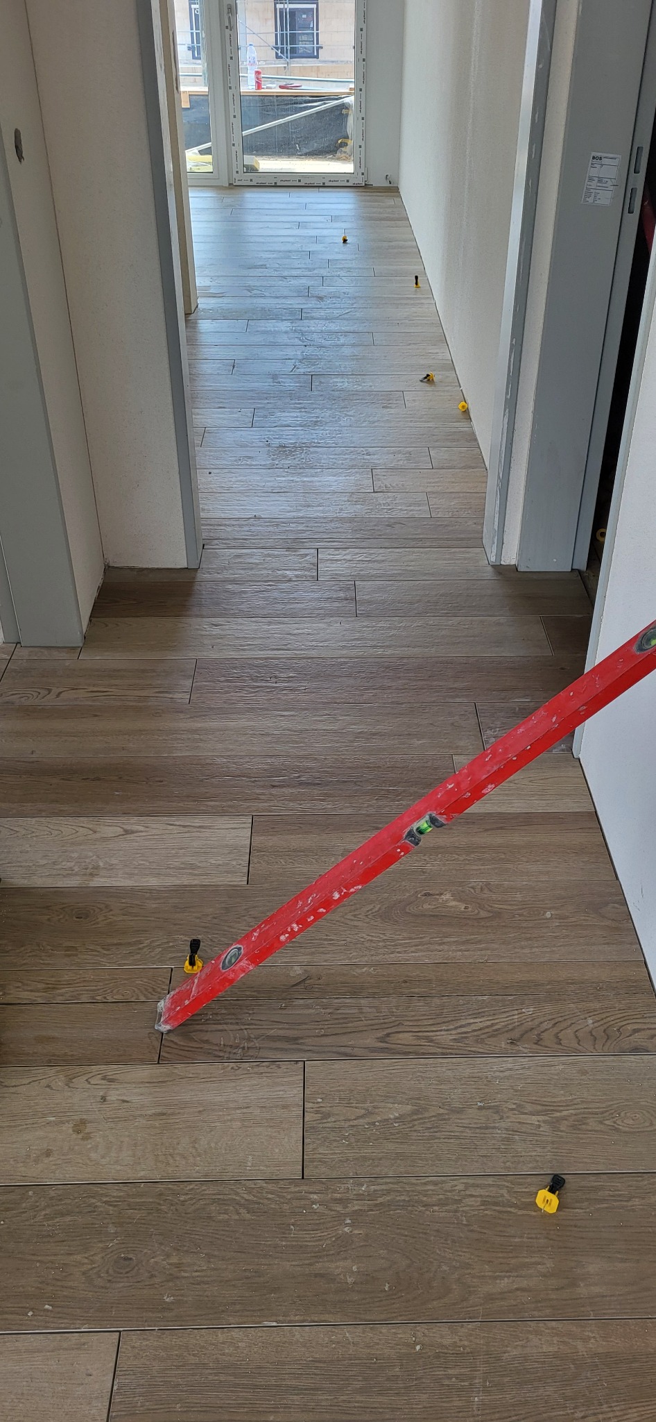 Ułożona podłoga z płytek imitujących drewno, z widocznym poziomem i elementami do poziomowania. Jasne wnętrze z oknem w tle.