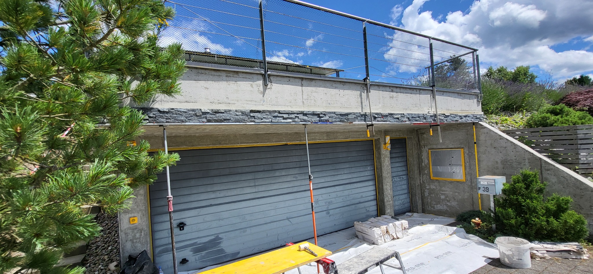 Montaż kamienia dekoracyjnego na elewacji garażu z widocznymi rusztowaniami i przygotowanym podłożem. Prace wykończeniowe w toku, widok na ogród i błękitne niebo.