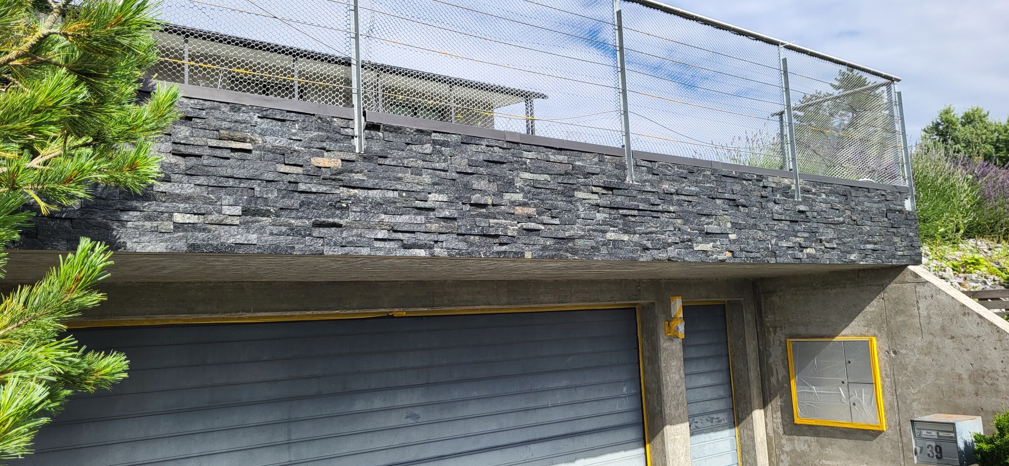 Elewacja balkonu wykończona czarnym kamieniem łupanym, zabezpieczona siatką. Widok z dołu na garaż z szarą bramą rolowaną i skrzynką pocztową.