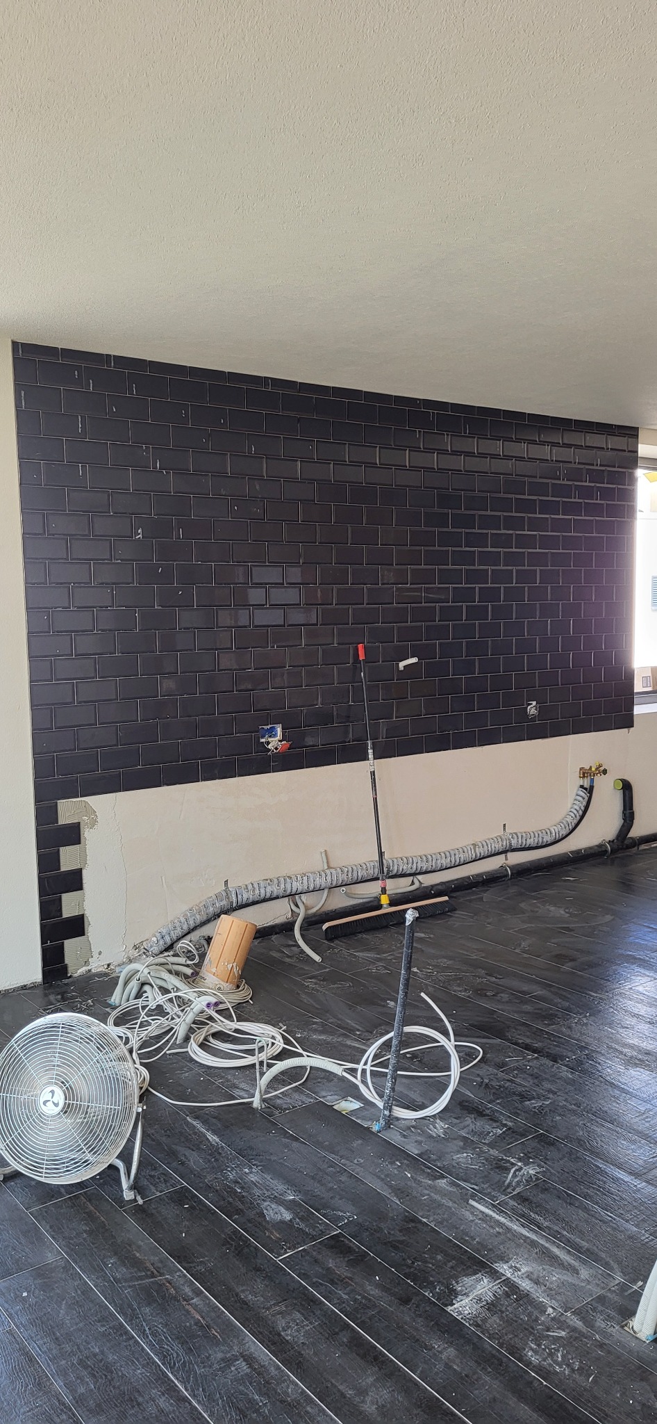 Częściowo wykończona ściana z czarnych płytek w kuchni, widoczne przewody i narzędzia glazurnicze. Podłoga wyłożona ciemnymi panelami imitującymi drewno.