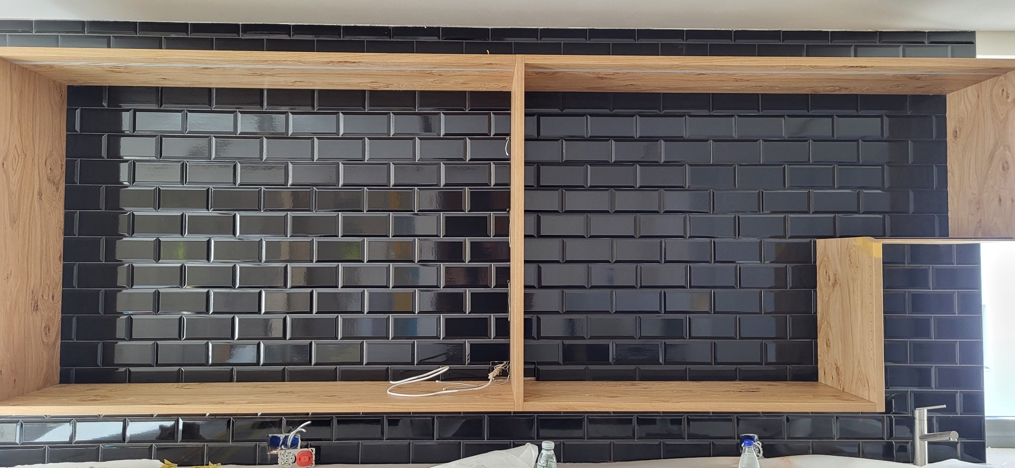 Błyszczące, czarne płytki typu metro na ścianie, z drewnianymi półkami bez frontów. Widoczne przewody elektryczne i fragment blatu z akcesoriami.