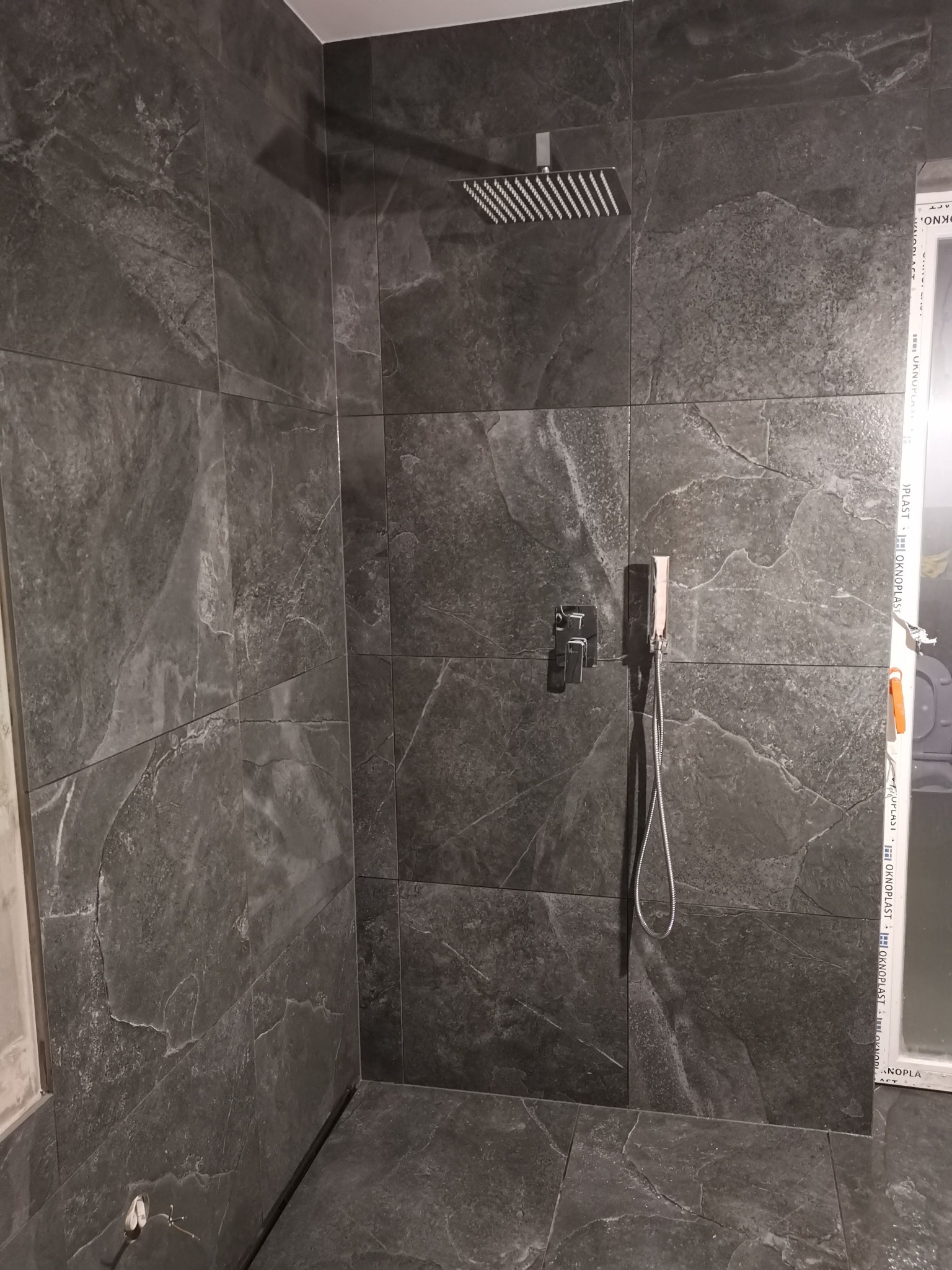 Nowoczesna kabina prysznicowa wyłożona ciemnymi, marmurkowymi płytkami. Widoczna deszczownica, bateria i okno z profilem PCV. Minimalistyczny design.