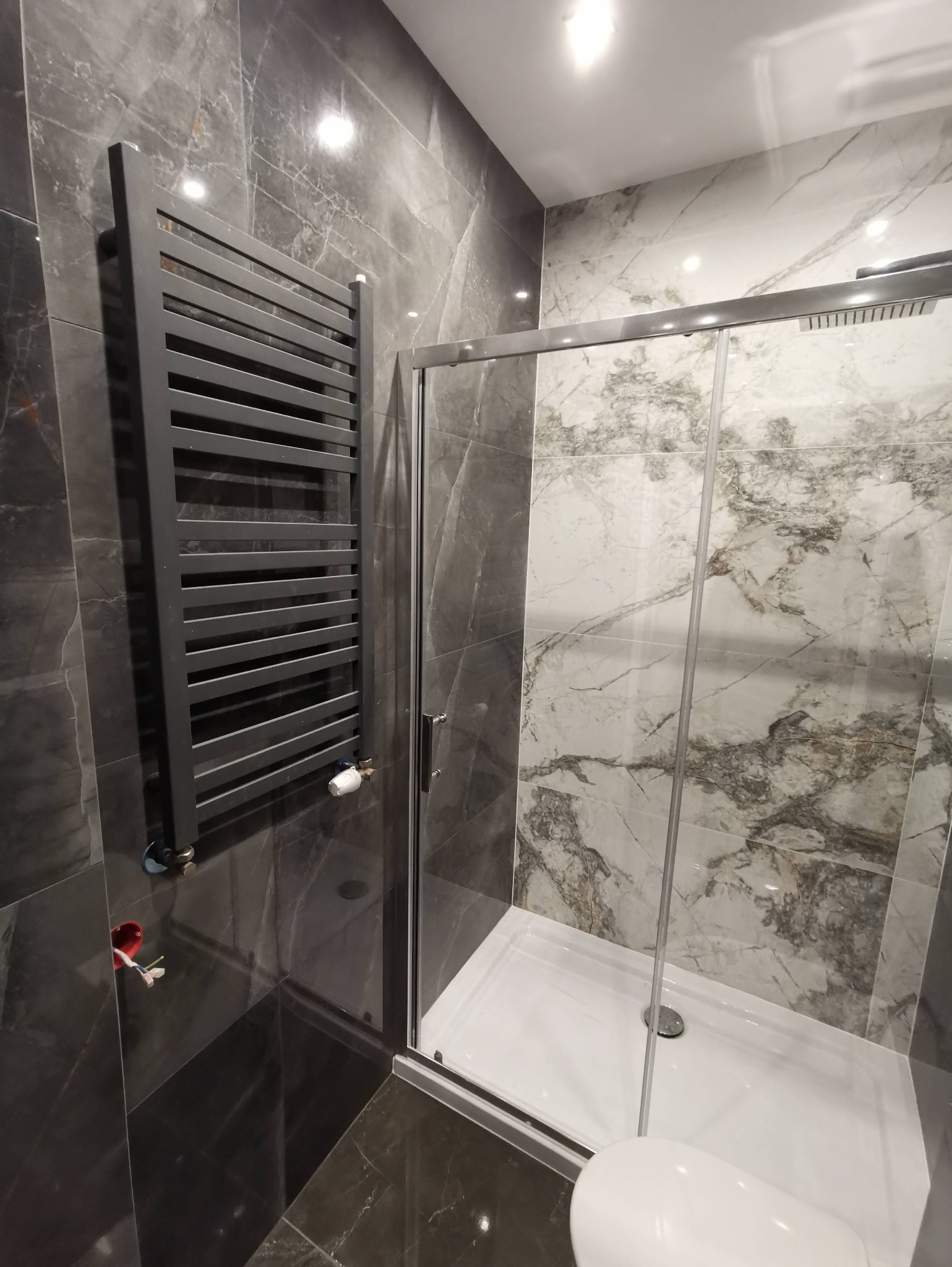 Nowoczesna łazienka z kabiną prysznicową i grzejnikiem drabinkowym na tle marmurowych płytek w odcieniach szarości. Widoczna instalacja elektryczna.