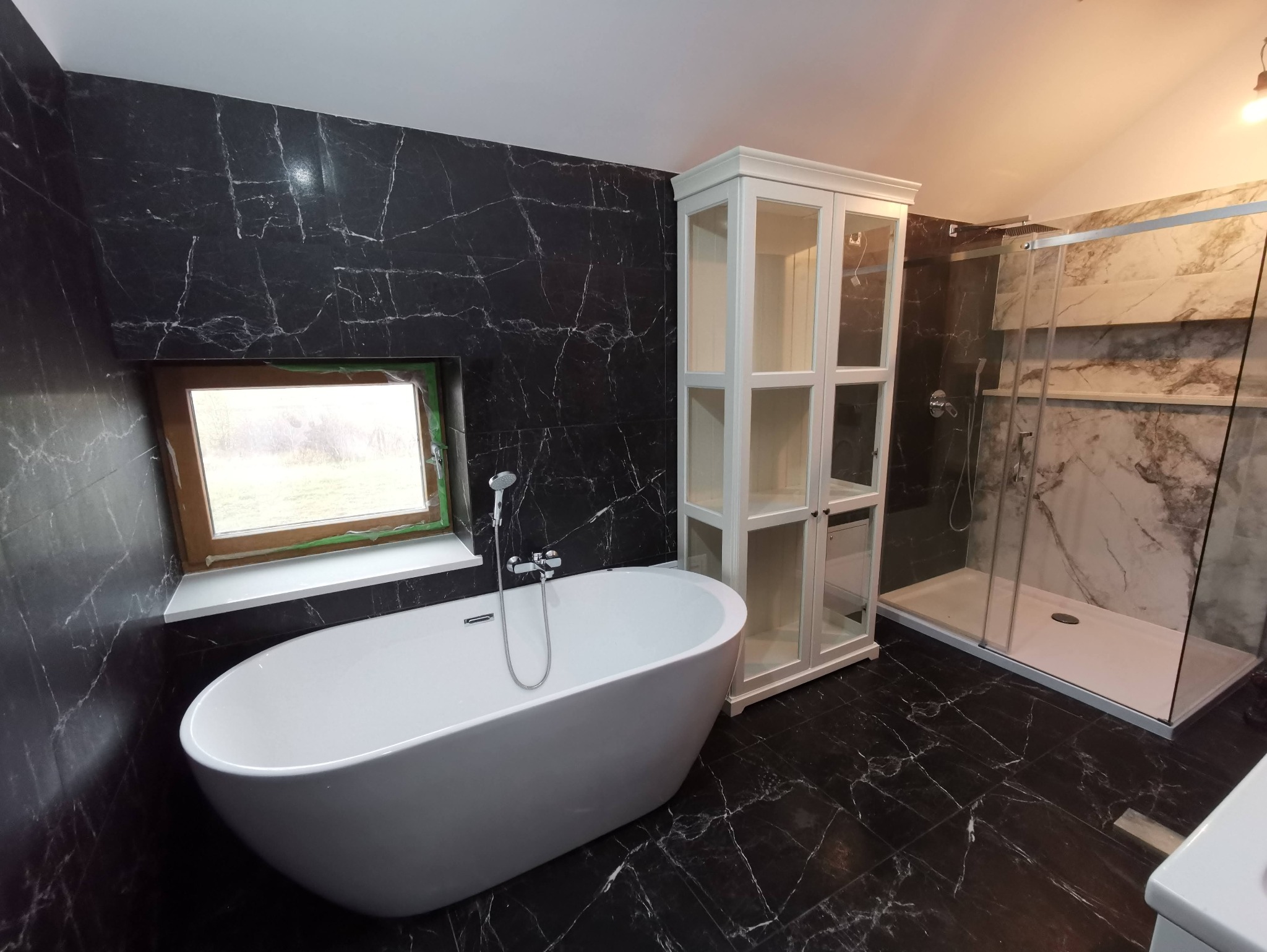 Elegancka łazienka z czarnymi marmurowymi płytkami, wolnostojącą białą wanną, kabiną prysznicową i białą szafką. Okno z drewnianą ramą dodaje naturalnego światła.
