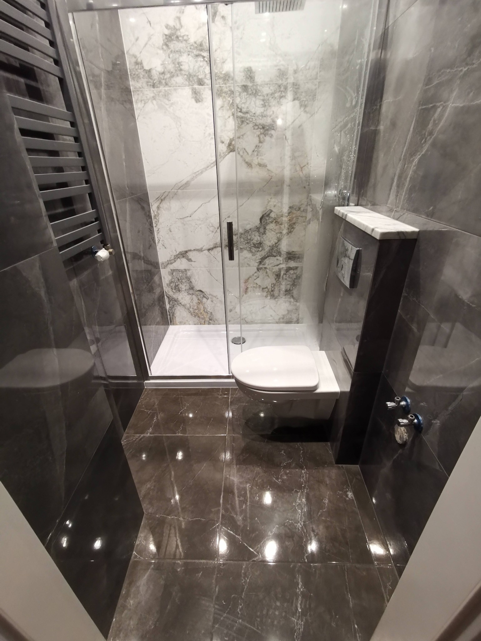 Nowoczesna łazienka z kabiną prysznicową typu walk-in, marmurowymi płytkami w odcieniach szarości i białym, podwieszanym WC. Widoczny grzejnik drabinkowy.