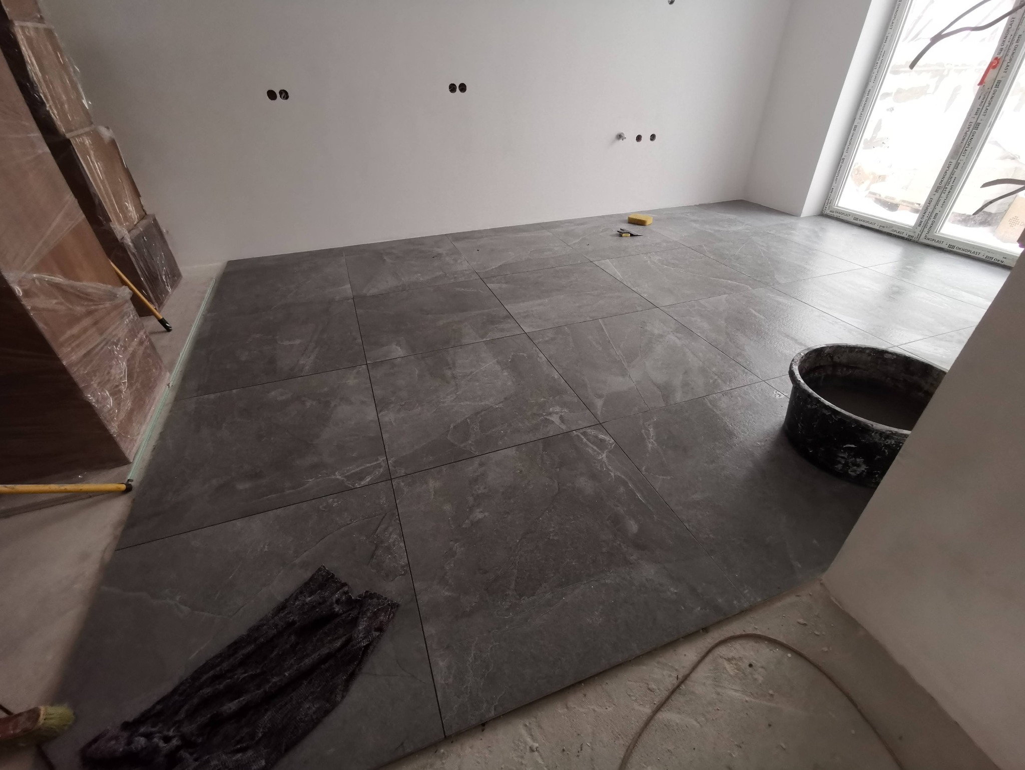 Ułożone, szare płytki podłogowe z efektem marmuru w jasnym pomieszczeniu. Widoczne narzędzia glazurnicze: wiadro z zaprawą, gąbka i szmatka. Świeżo położona podłoga.