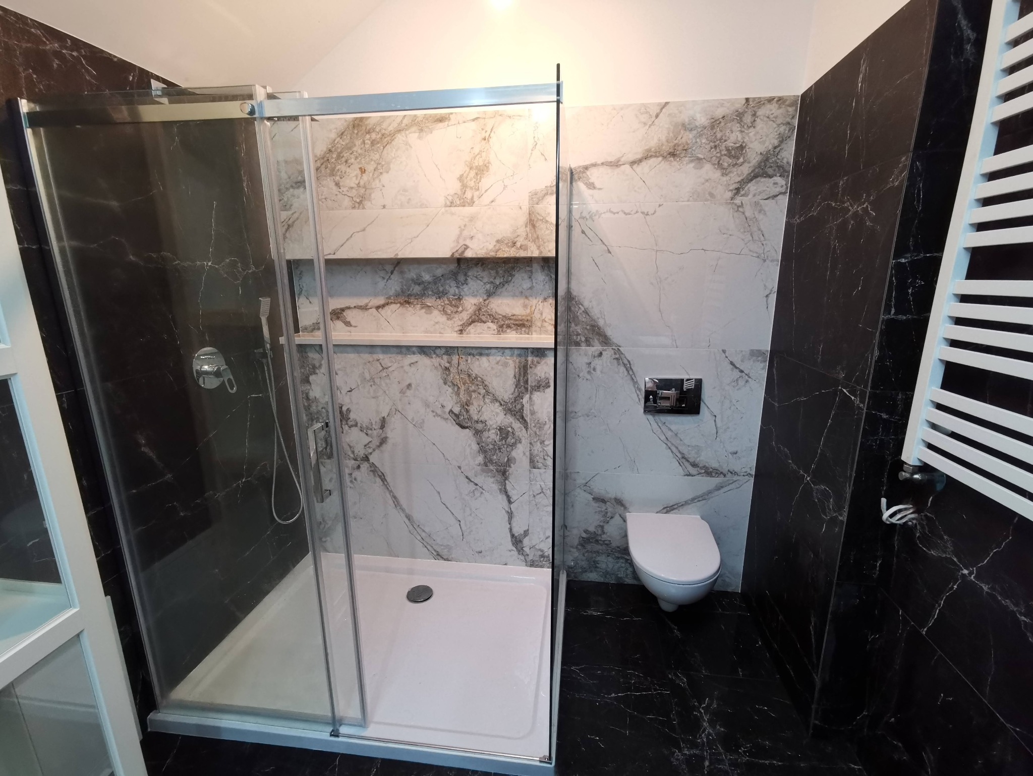 Nowoczesna łazienka z kabiną prysznicową typu walk-in, wykończona płytkami imitującymi marmur w odcieniach bieli i czerni. Widoczny grzejnik drabinkowy i podwieszana toaleta.