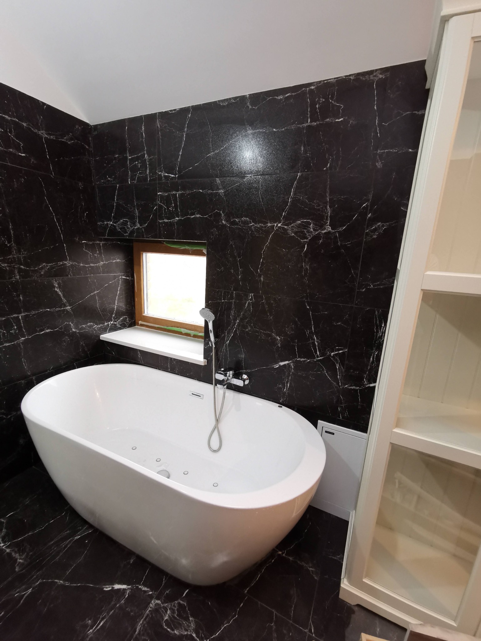 Nowoczesna łazienka z wanną wolnostojącą na tle czarnych płytek imitujących marmur. Małe okno z drewnianą ramą i biały regał z półkami po prawej.