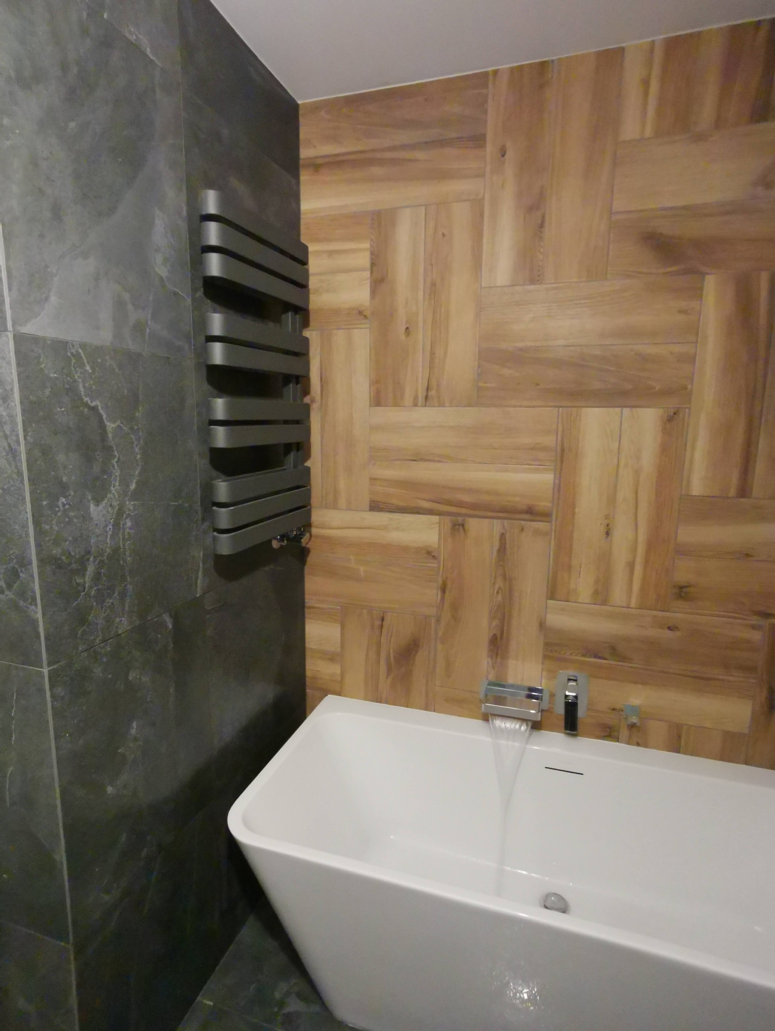 Nowoczesna łazienka z wanną, grzejnikiem drabinkowym na tle ściany z szarych płytek imitujących kamień i drewnopodobnych płytek ułożonych w jodełkę.