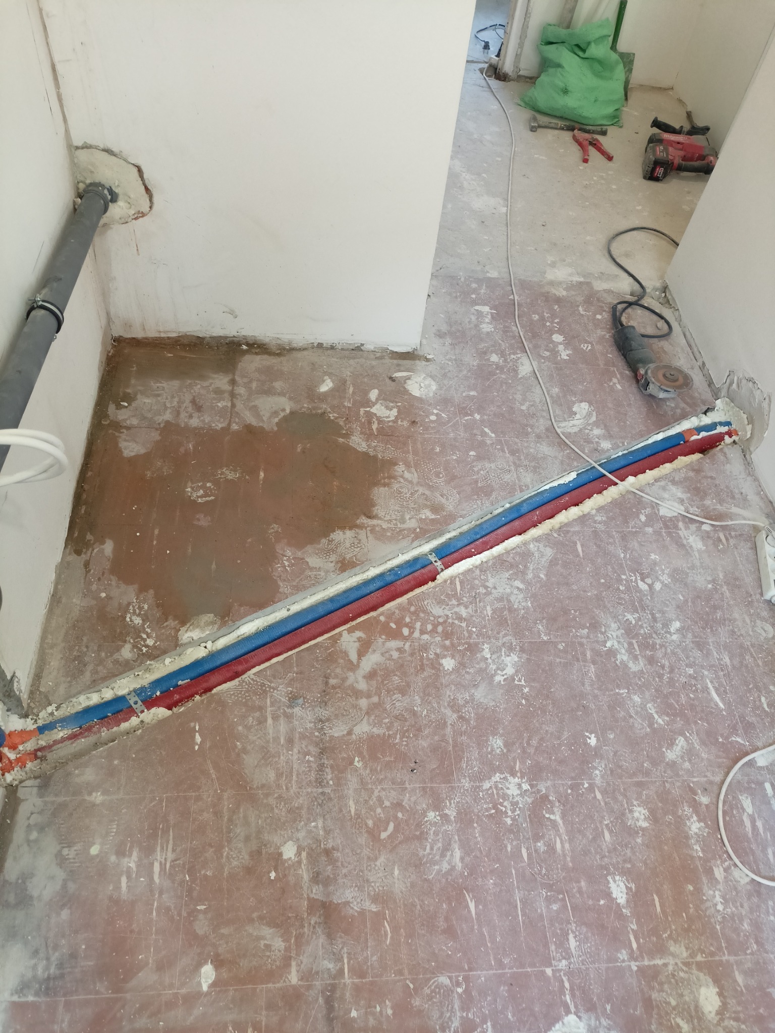 Instalacja rur grzewczych w podłodze, zabezpieczonych pianką montażową. Widoczne narzędzia i materiały budowlane. Ujęcie z góry, w trakcie prac instalacyjnych.