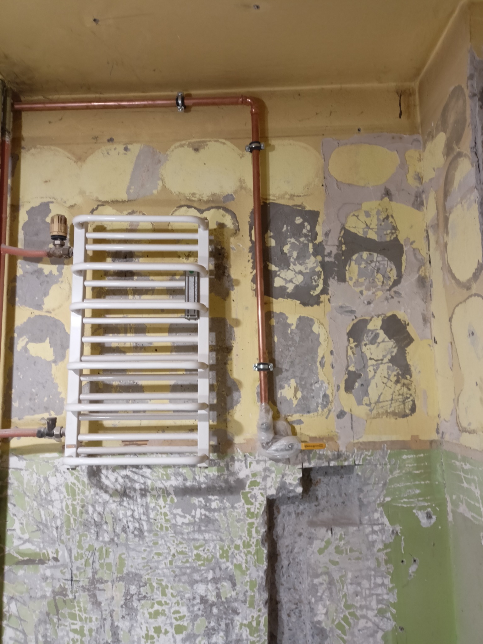 Biały grzejnik drabinkowy zamontowany na ścianie z widocznymi śladami po remoncie, miedziane rury instalacji grzewczej.