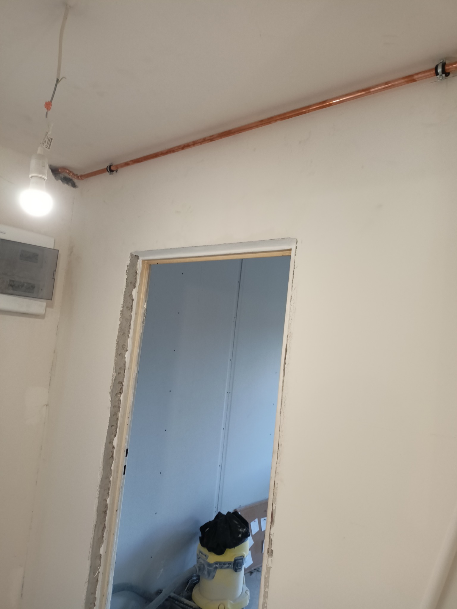 Miedziana rura instalacji biegnąca wzdłuż sufitu w surowym wnętrzu. Widoczna żarówka na kablu i odkurzacz przemysłowy. Ściany w trakcie wykończenia.