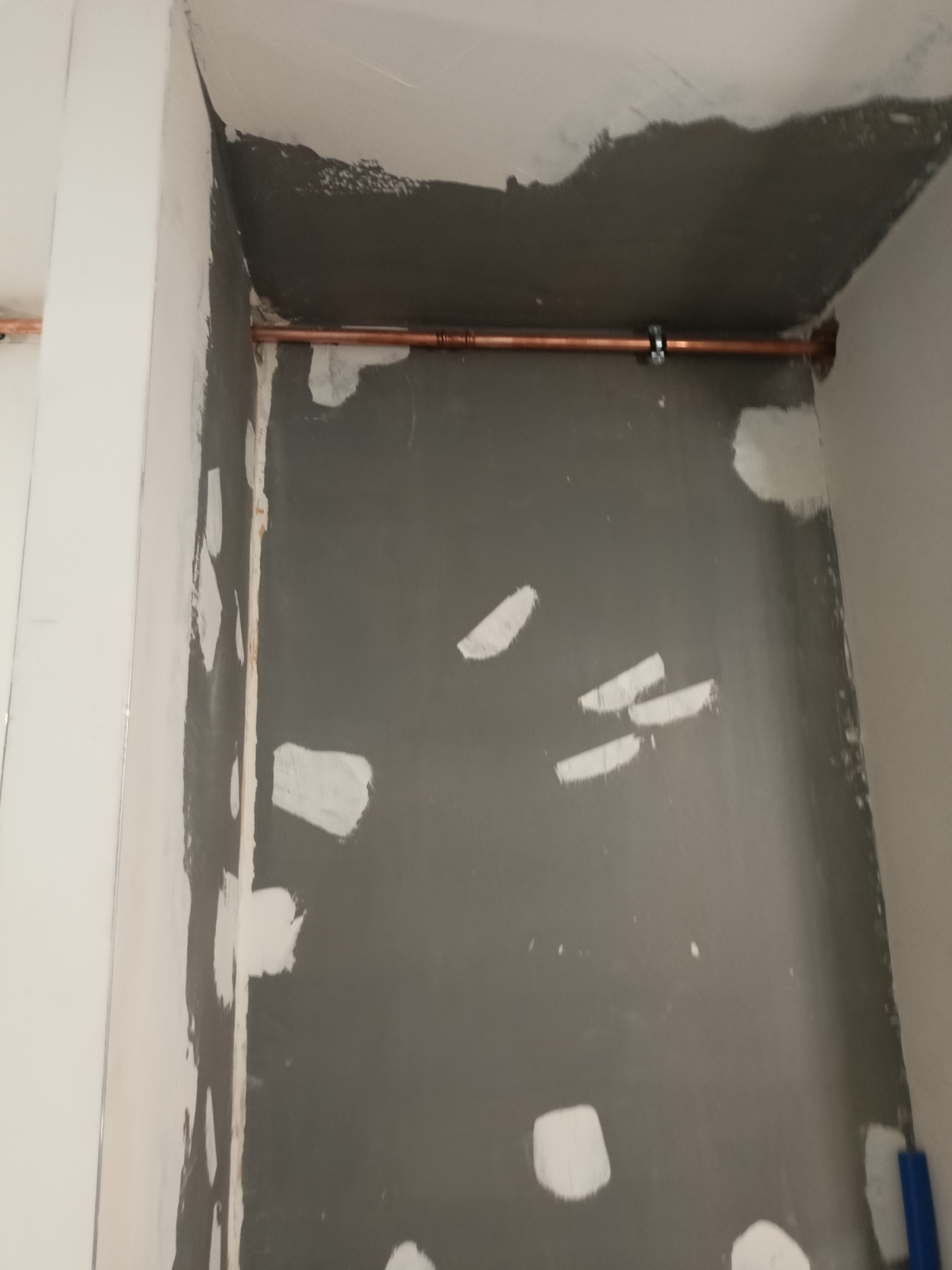 Miedziana rura instalacyjna zamocowana do surowej, szarej ściany z widocznymi łatami gipsowymi. Fragment białej ściany po lewej stronie. Ujęcie z dołu.
