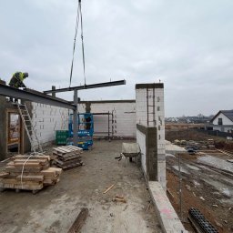 Budomad Michał Domagała - Montaż stalowej belki dźwigiem na budowie domu z białych bloczków. Widoczny pracownik na drabinie, podnośnik i palety z materiałami. Tło: zabudowa jednorodzinna.