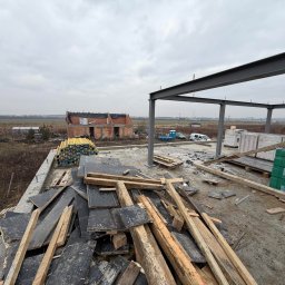 Budomad Michał Domagała - Budowa domu w trakcie: stalowa konstrukcja, palety z bloczkami silikatowymi H+H, drewniane deski i w tle budynek w stanie surowym otwartym, pochmurne niebo.