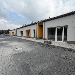 Budomad Michał Domagała - Parterowy budynek z szarymi ścianami i żółtymi akcentami okiennymi, widoczny z zewnątrz, z nową kostką brukową na podjeździe i elementami wykończeniowymi.